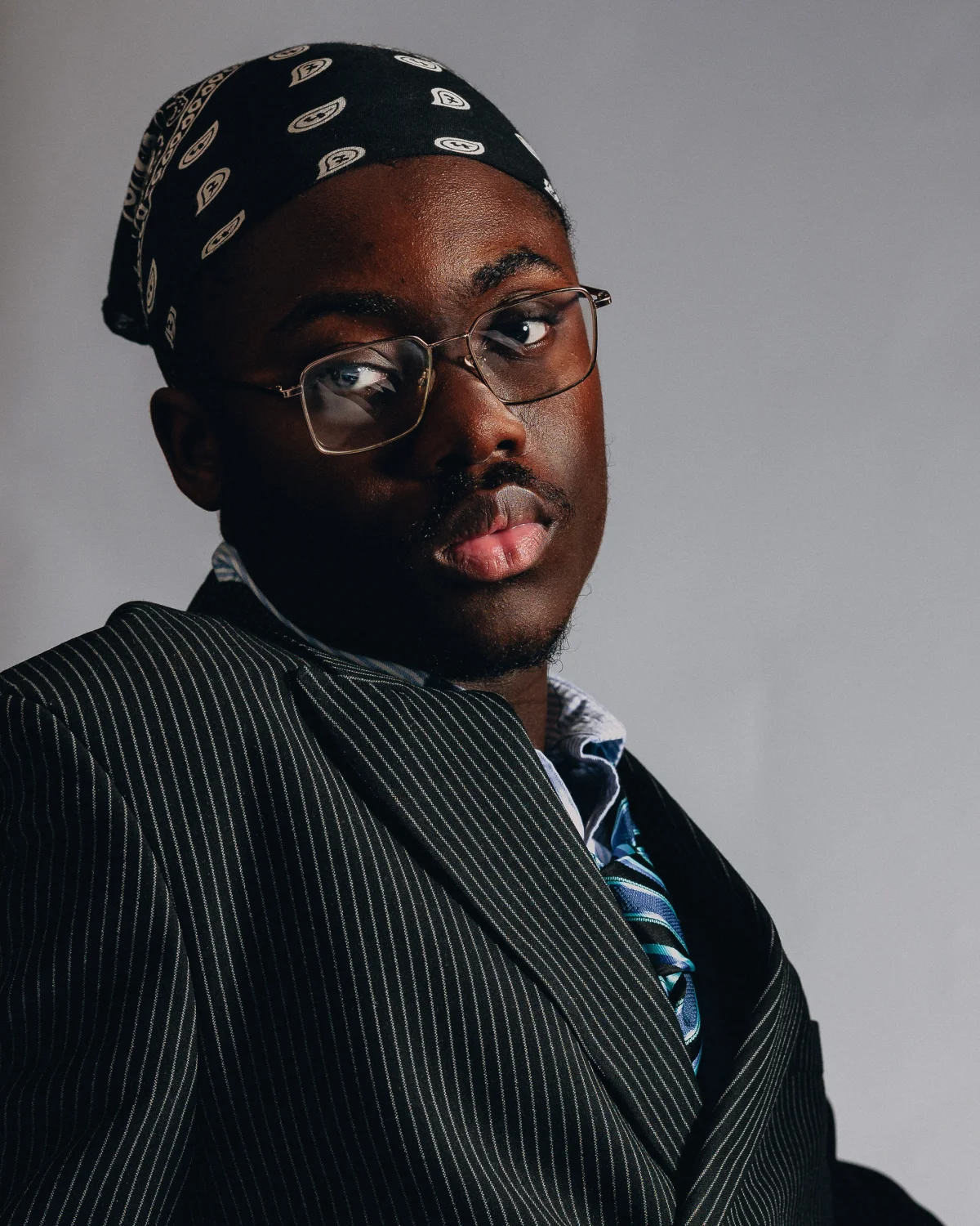 Close-up portrait of a man wearing glasses, a black patterned headwrap, and a pinstripe suit against a grey background.