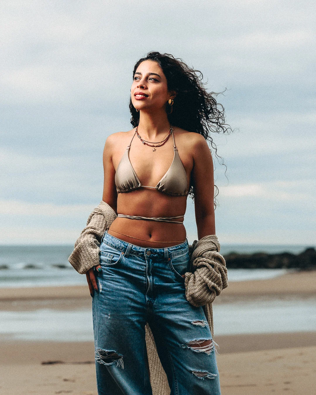 A young woman with curly hair posing on a beach wearing a gold bikini top, ripped blue jeans, and a beige cardigan.