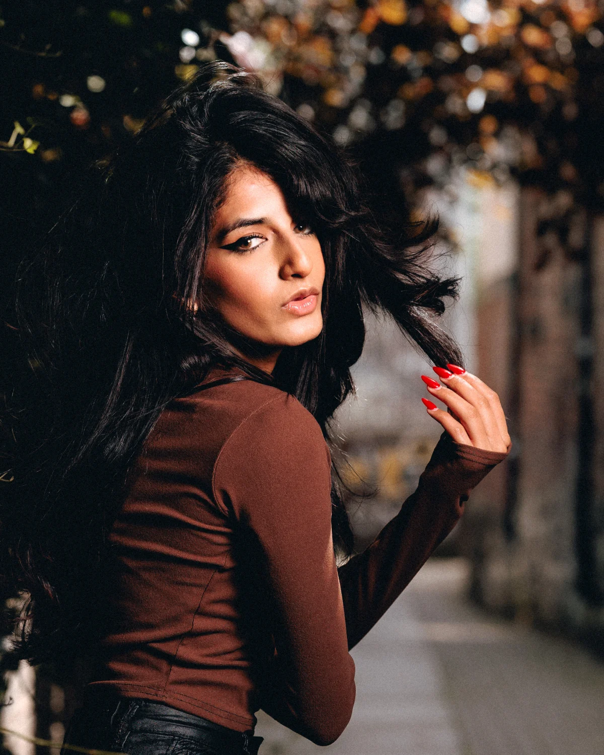 A woman with long dark hair and red nails posing outdoors in a brown top, looking over her shoulder.