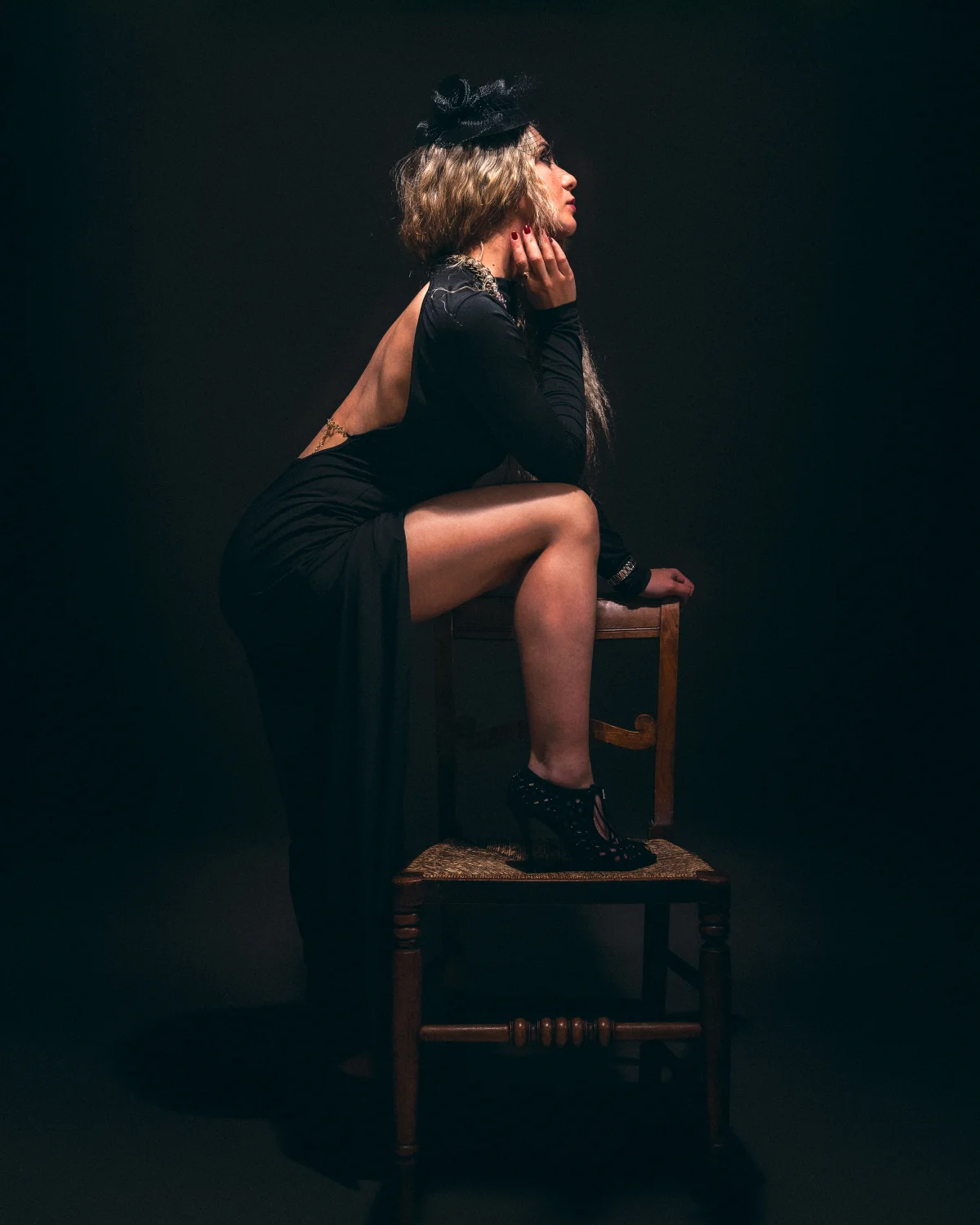 A woman in an elegant black evening gown and fascinator hat poses on a wooden chair against a dark background.