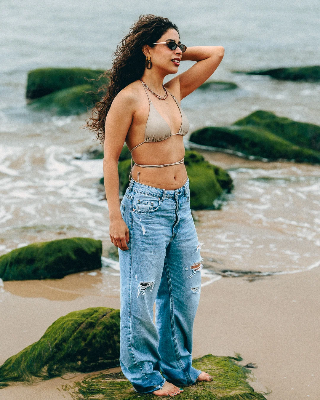 Woman with curly hair in a beige bikini top and ripped jeans posing on a rocky beach.