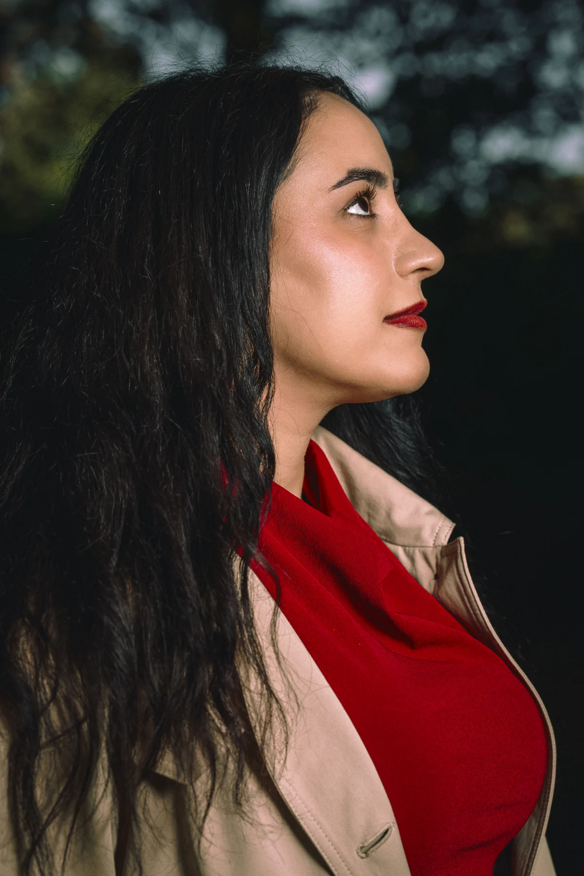 Side profile portrait of a woman with long dark hair, red lipstick, wearing a beige coat and red top outdoors.