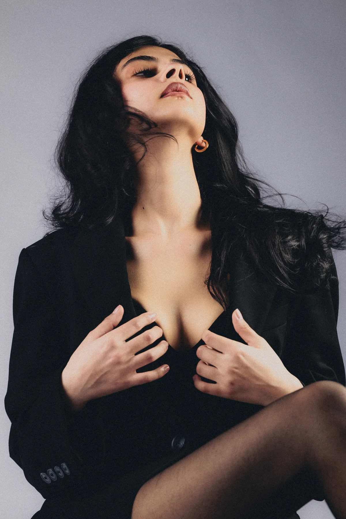 Woman with long dark hair wearing an open black blazer, posing with head tilted back against a grey background.