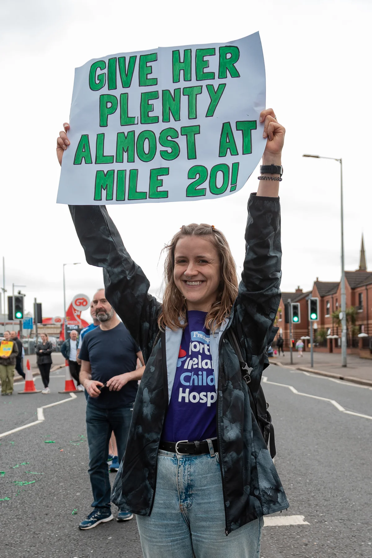 A smiling woman outdoors holding a sign that reads 'GIVE HER PLENTY ALMOST AT MILE 20!' during a race event.