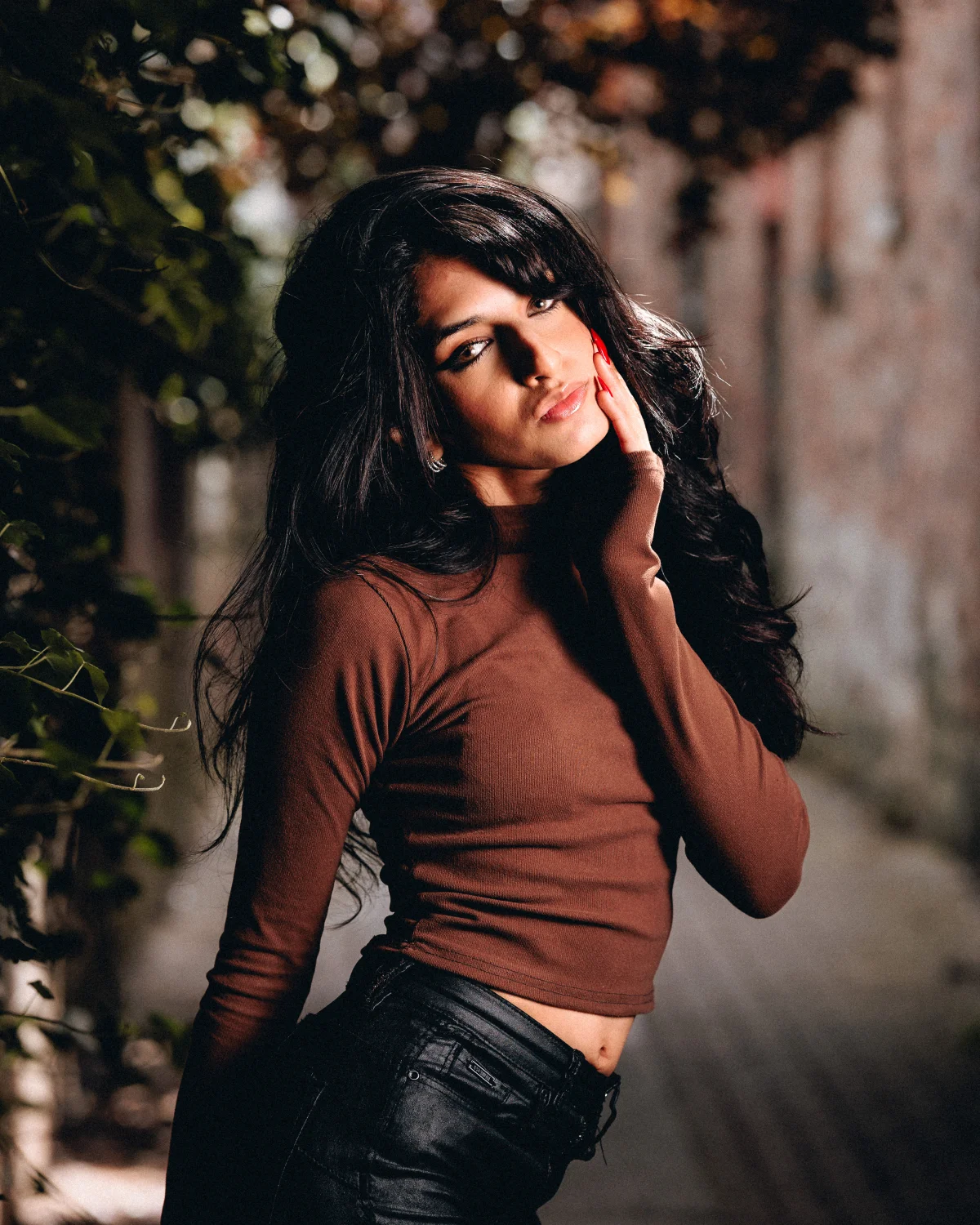 A young woman with long dark hair in a brown turtleneck and black leather pants posing outdoors.
