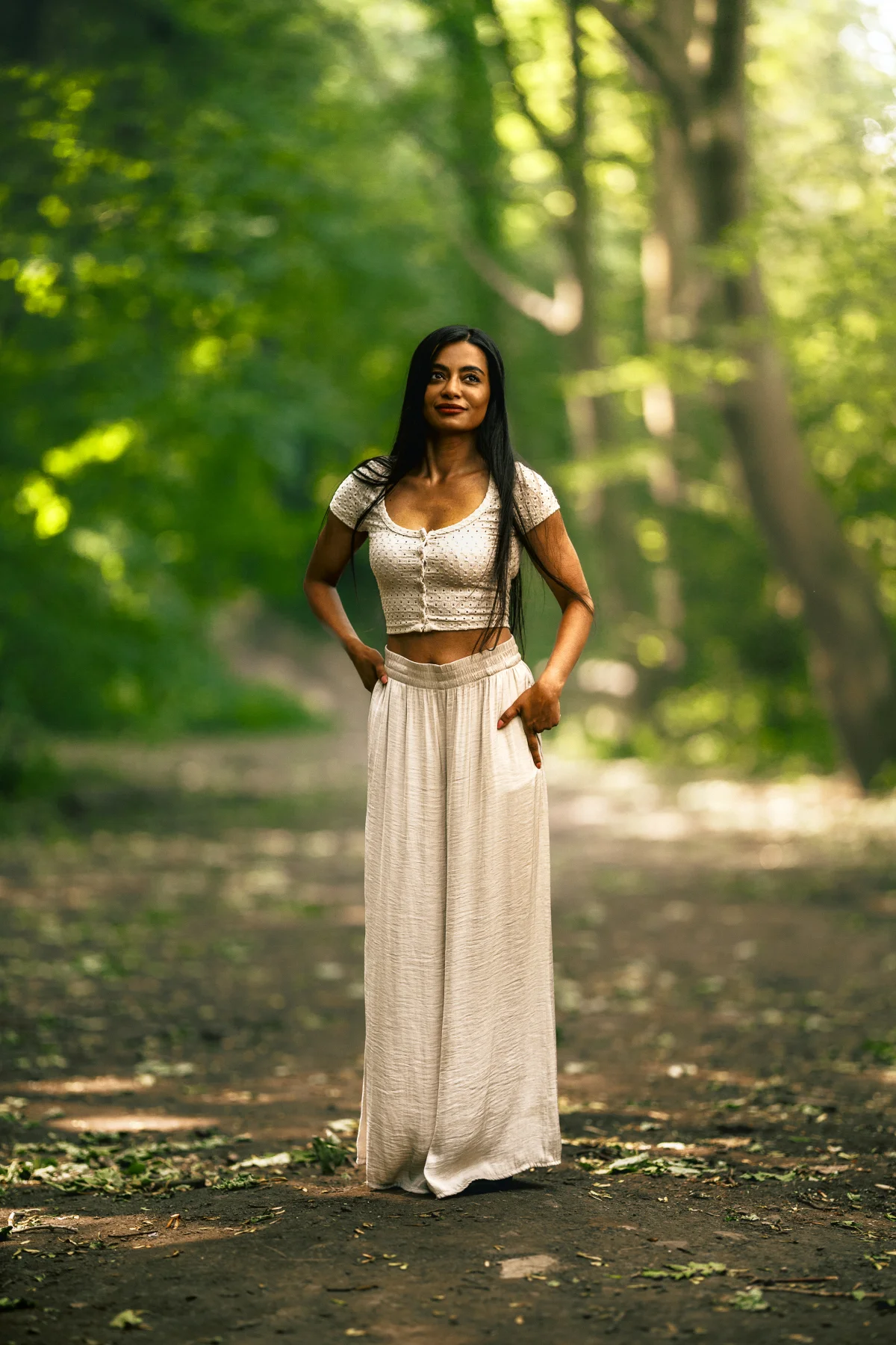 A woman with long dark hair wearing a white crop top and matching long skirt posing on a forest path.