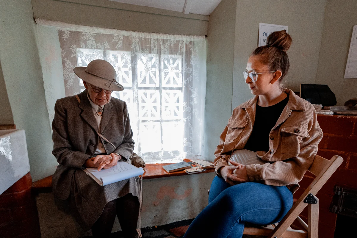 An older woman in vintage attire and a younger woman in modern clothing sitting together indoors.