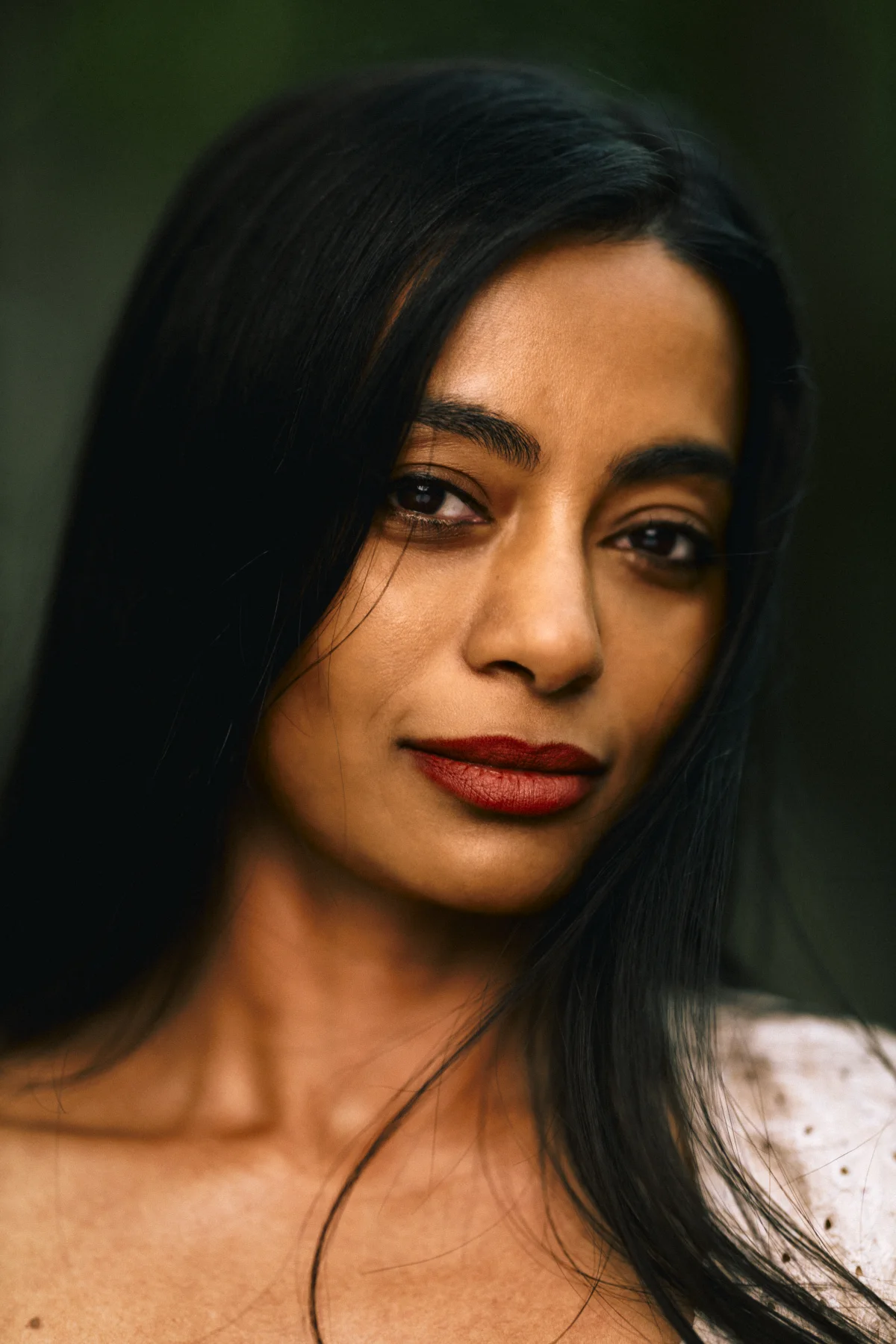 Close-up portrait of a woman with long dark hair and red lipstick against a soft, blurred green background.