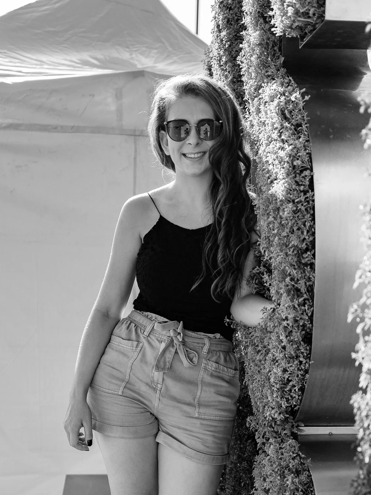 Black and white portrait of a smiling woman in sunglasses, black top, and denim shorts leaning against greenery.