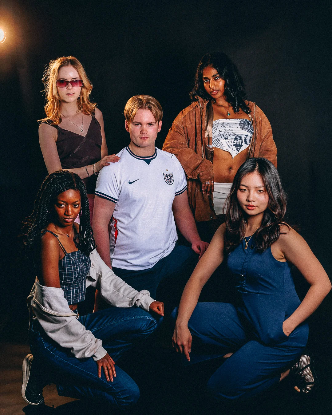 A group of five diverse young adults posing in a studio against a dark background, wearing trendy casual attire.
