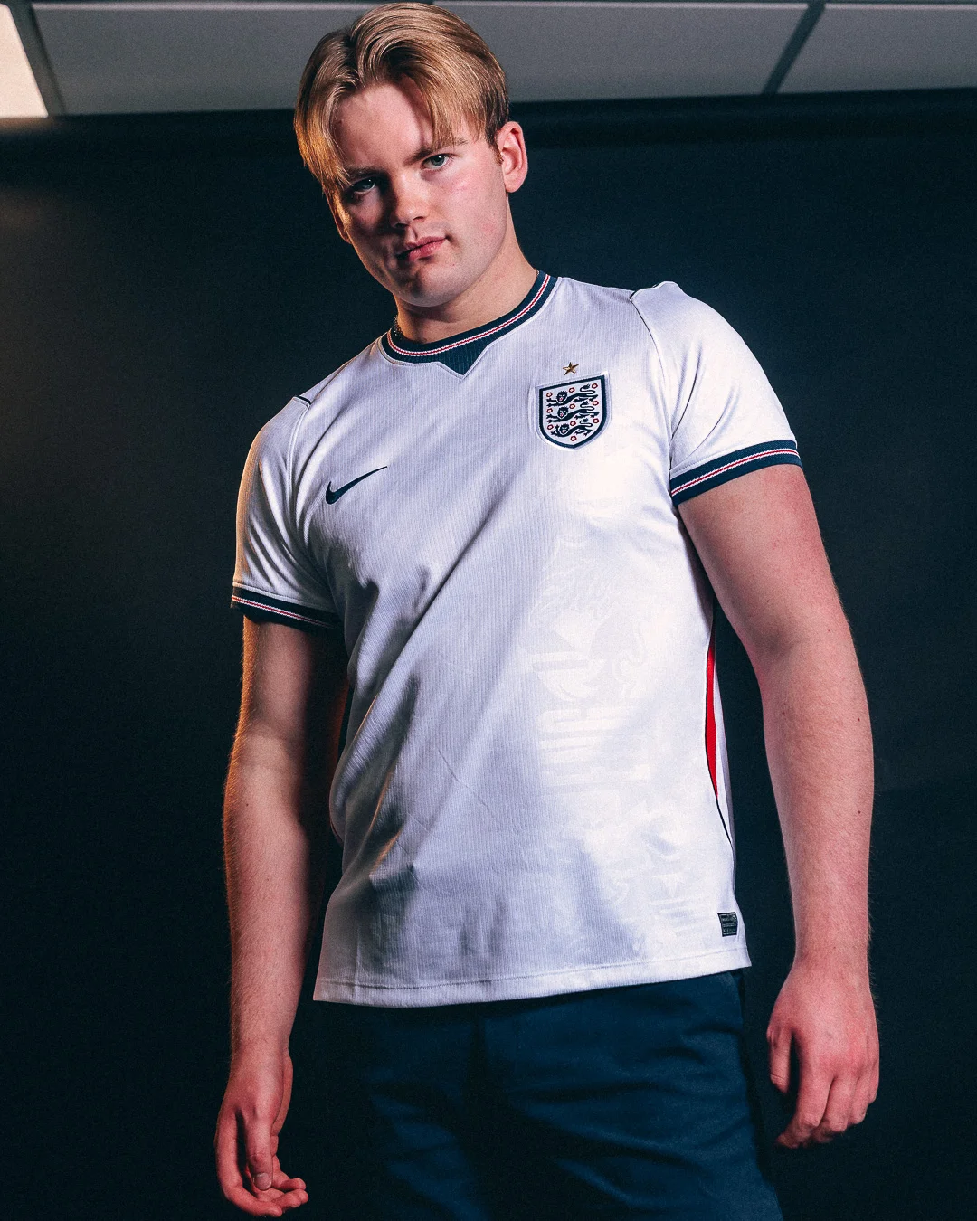 A young man with blonde hair wearing an England football jersey posing against a dark background.