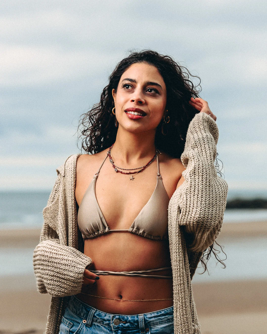 A woman with curly dark hair posing on a beach in a beige bikini top and an oversized knit cardigan.