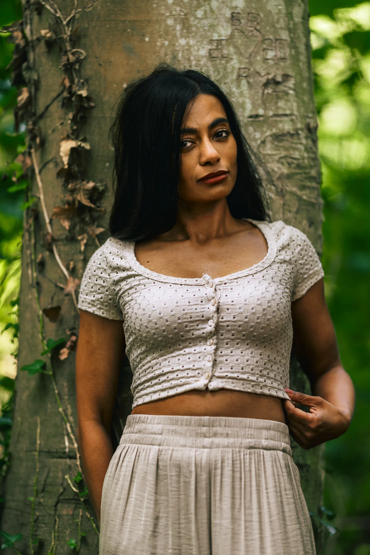 A woman with long dark hair wearing a white textured crop top, leaning against a large tree in a forest.