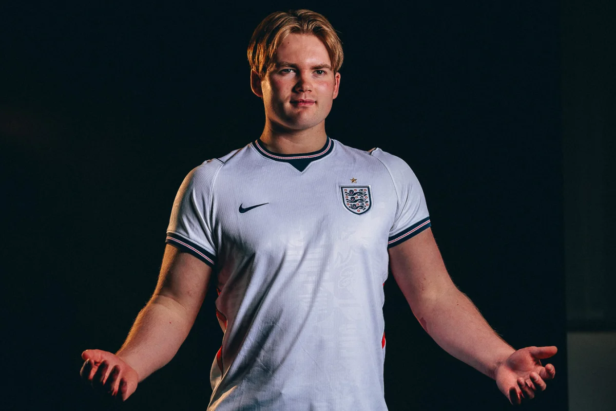 A man in an England football jersey posing against a dark background with dramatic studio lighting.