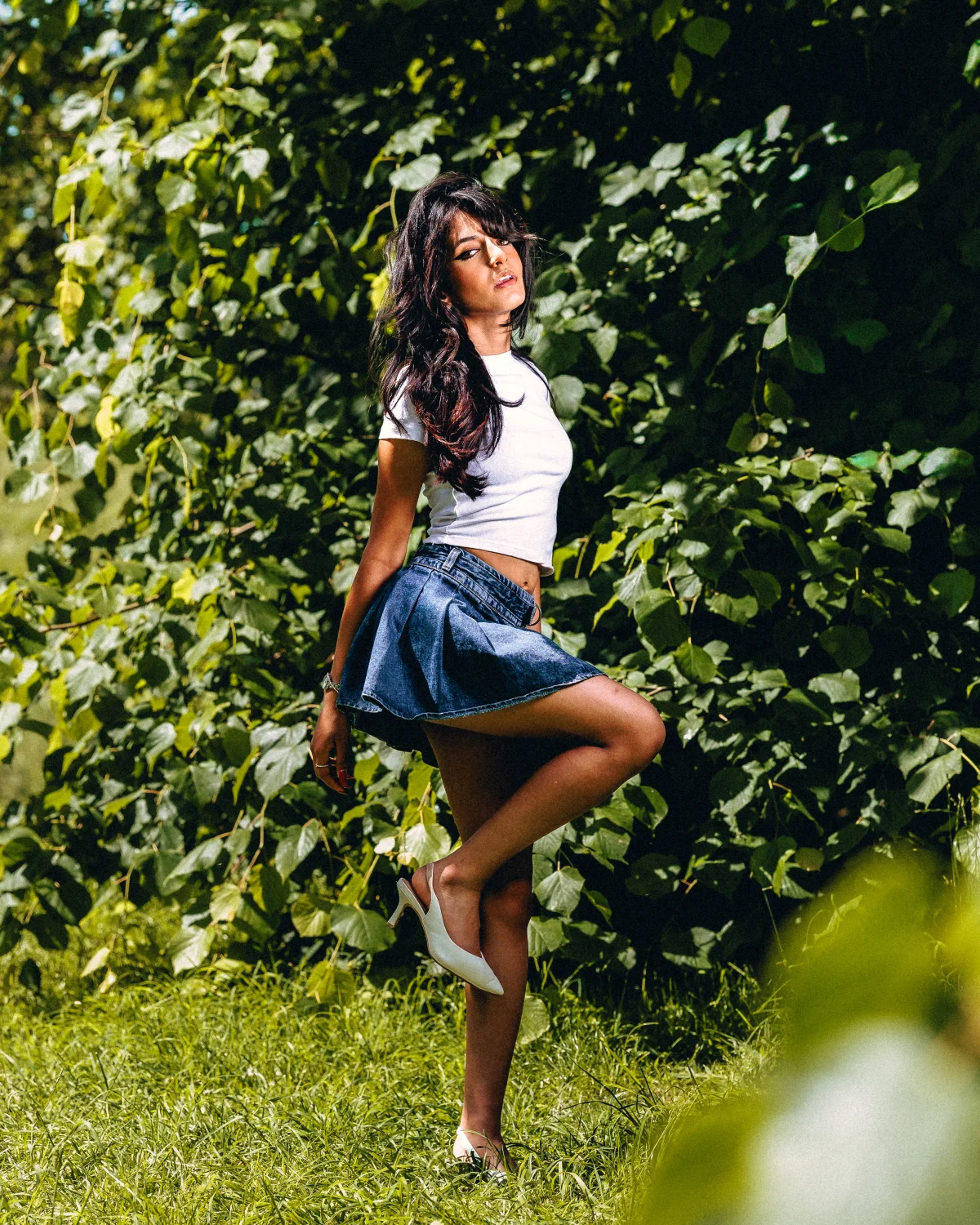 Full body shot of a woman in a white t-shirt and denim mini skirt posing against green foliage.