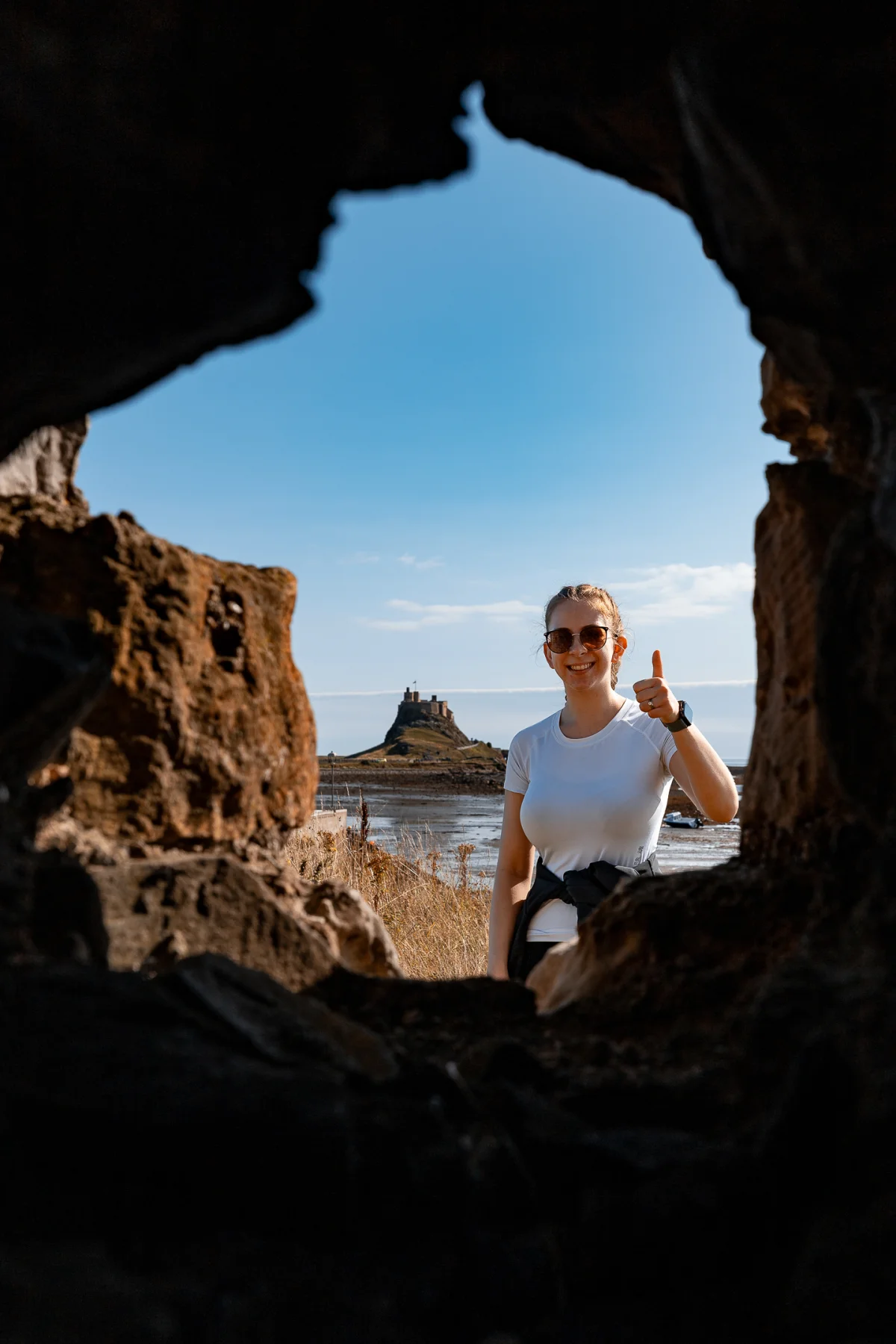 A woman in a white t-shirt giving a thumbs up, framed by dark rocks with a castle in the distance.