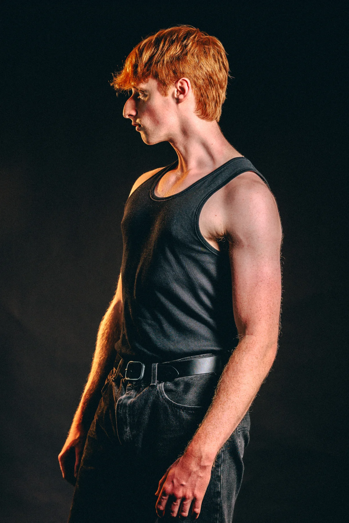 Profile portrait of a young man with red hair wearing a black tank top and jeans against a dark background.