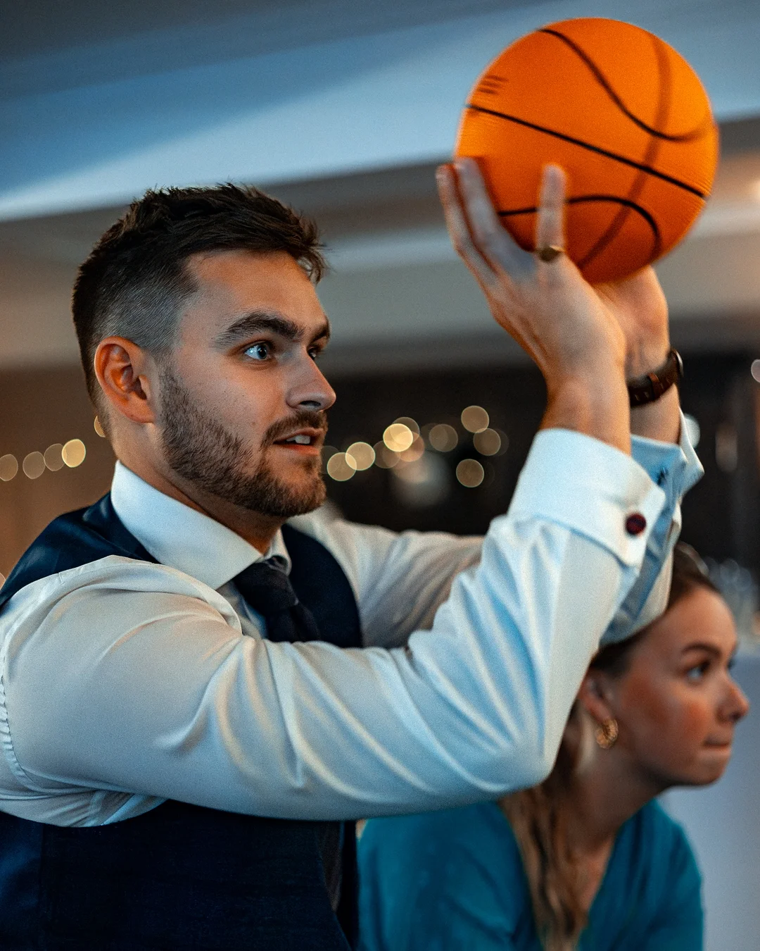 A man in a white shirt and navy vest holding an orange basketball aloft against a blurred indoor background.
