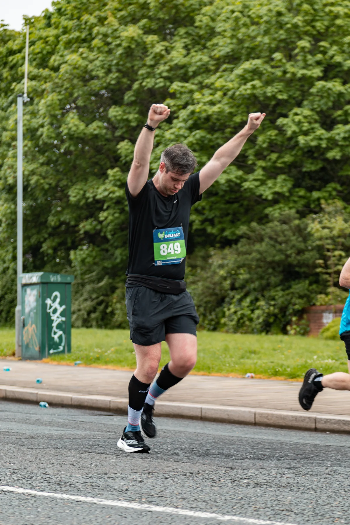 A male runner celebrating with raised arms after a race in an outdoor park setting.