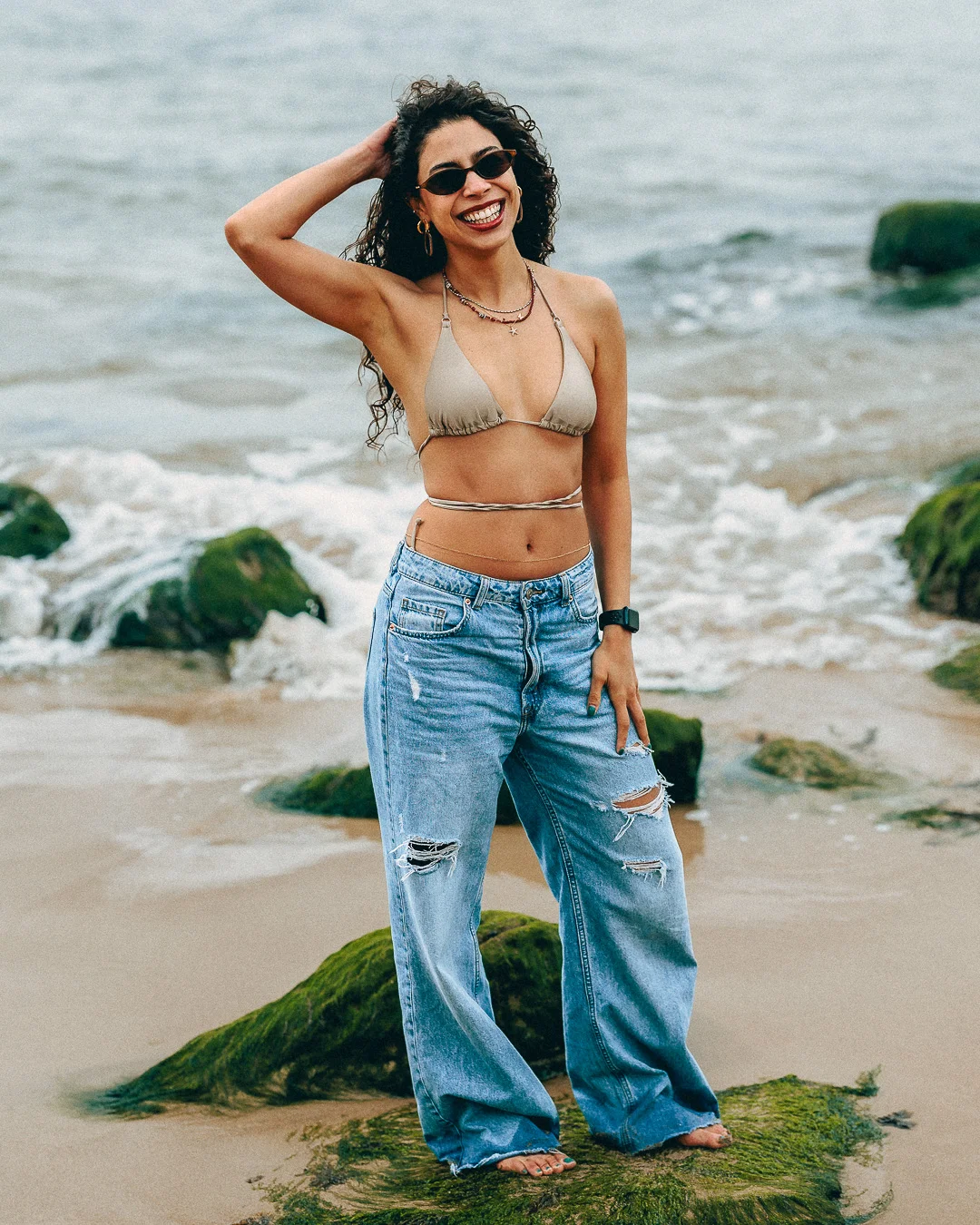 Smiling woman in a beige bikini top and ripped blue jeans posing on a beach with ocean waves.