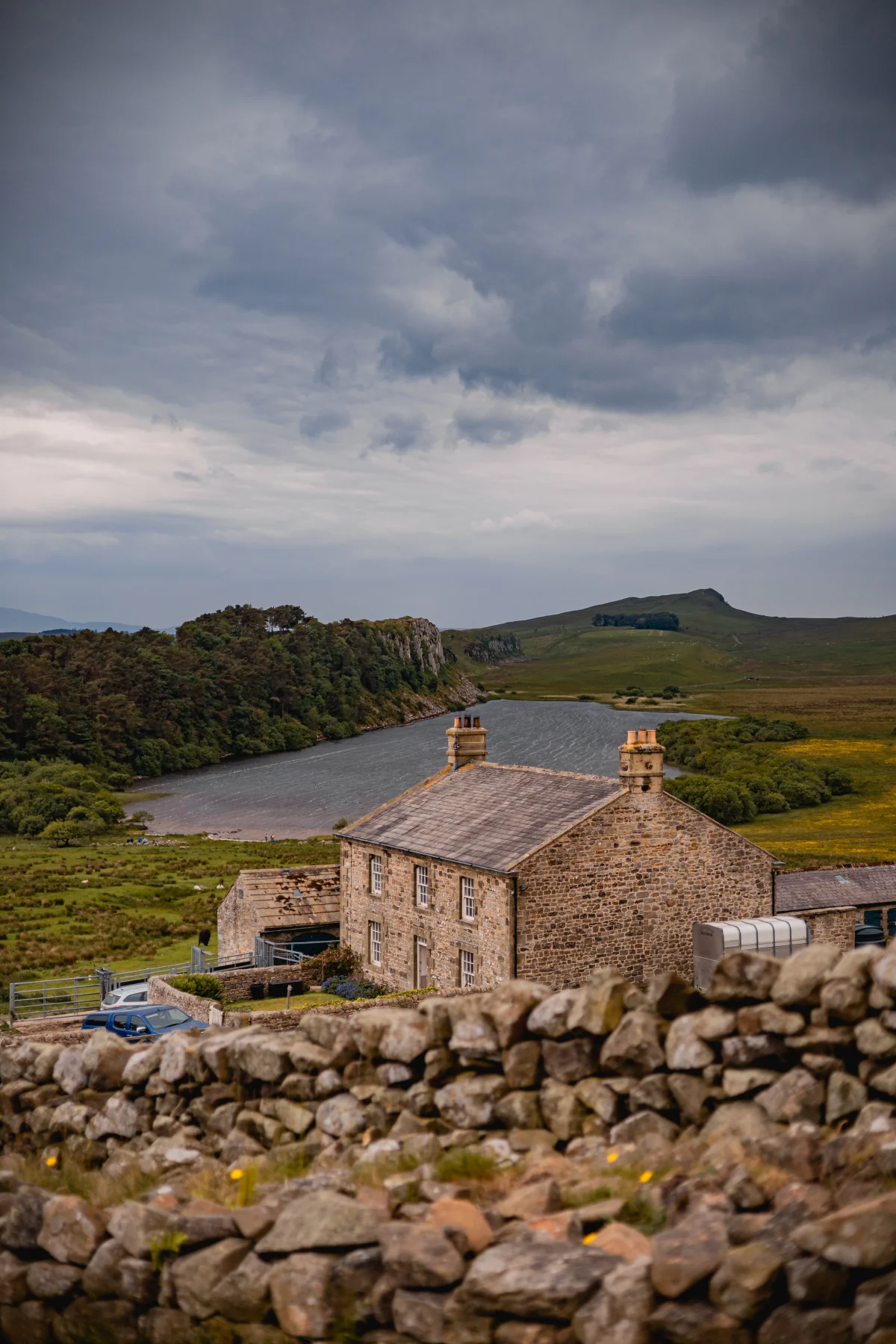 A rustic stone cottage by a serene lake under a moody, overcast sky in a hilly landscape.