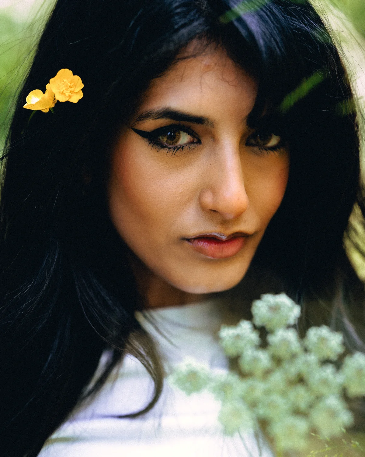 Close-up portrait of a woman with bold eyeliner and yellow flowers in her dark hair against a blurred green background.