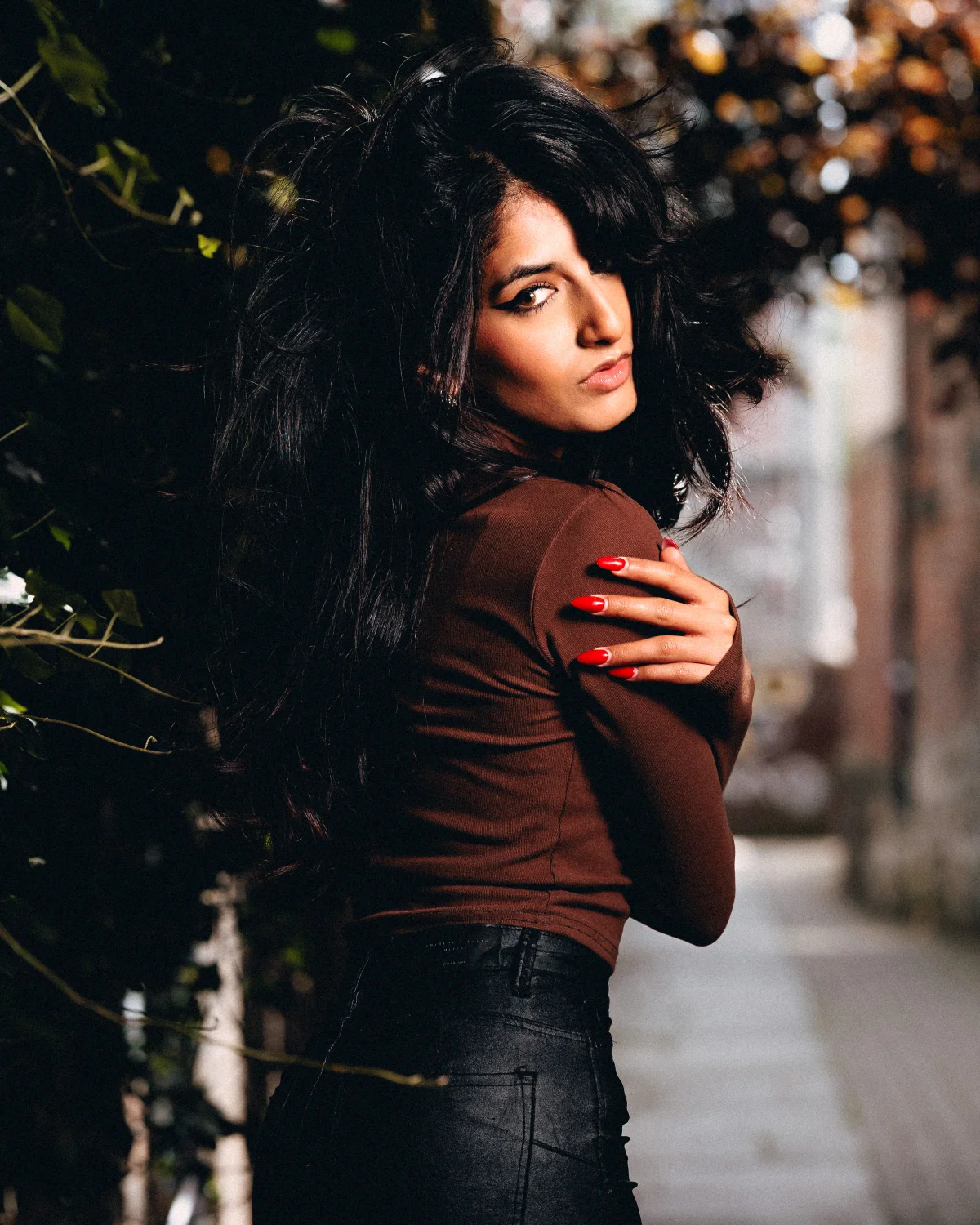 A woman with long dark hair in a brown turtleneck and black leather pants posing outdoors.