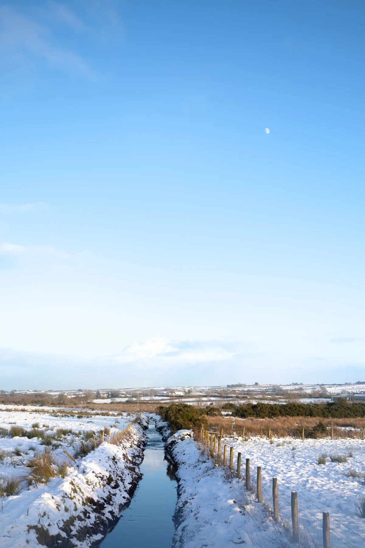 A narrow stream winding through a snowy field under a clear blue sky with a small moon.