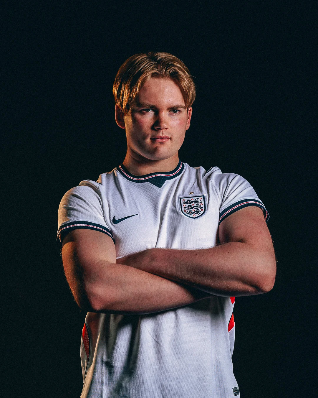 A young man with blond hair in an England football jersey, arms crossed against a dark background.