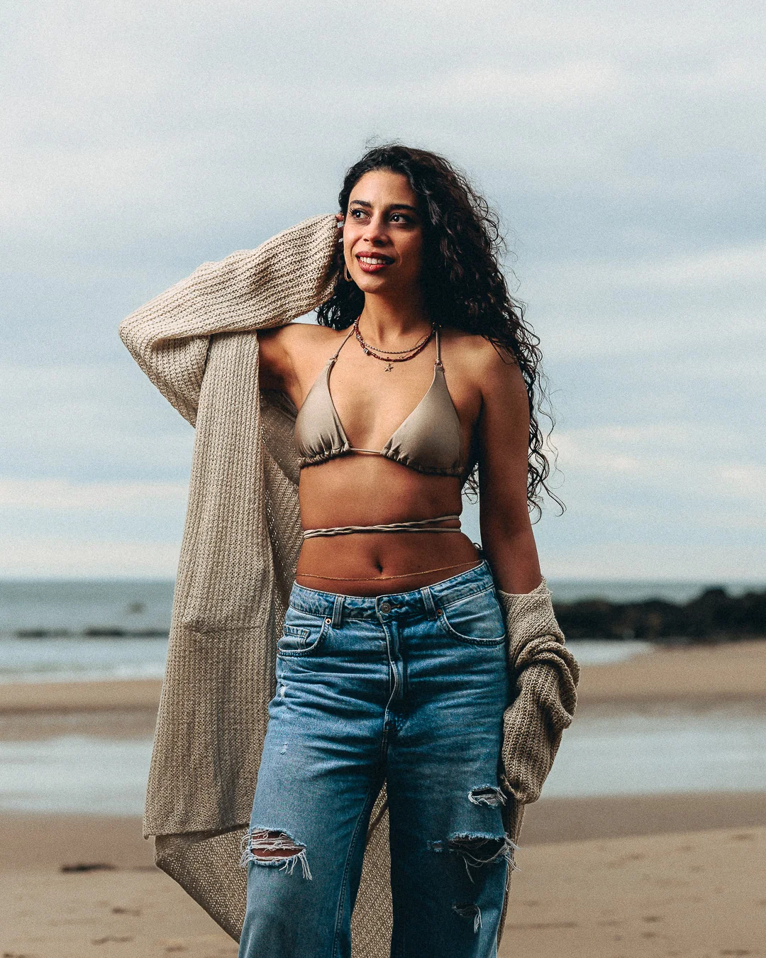 Woman with curly hair in a bikini top and ripped jeans wearing an oversized cardigan on a beach.