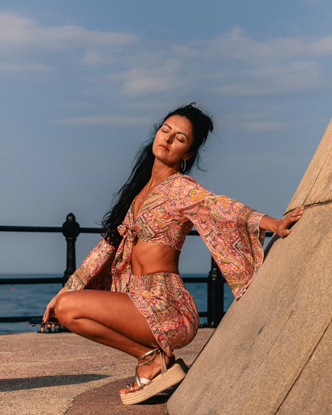 Woman in a pink patterned two-piece outfit posing by the sea with her eyes closed.
