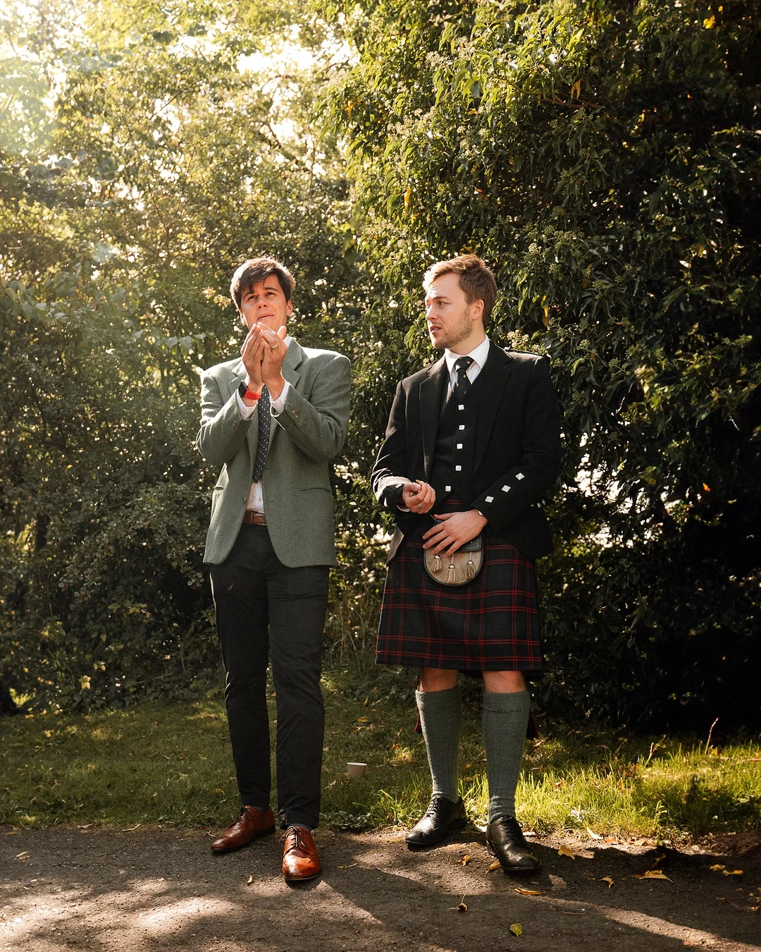 Two men in formal attire, one wearing a red tartan kilt, standing outdoors amidst lush green foliage.