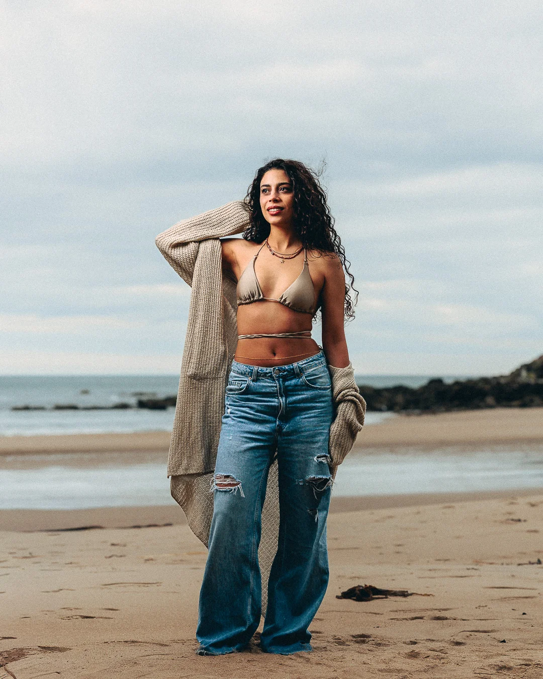 Woman with curly hair in a gold bikini top and wide-leg jeans posing on a beach with an oversized cardigan.