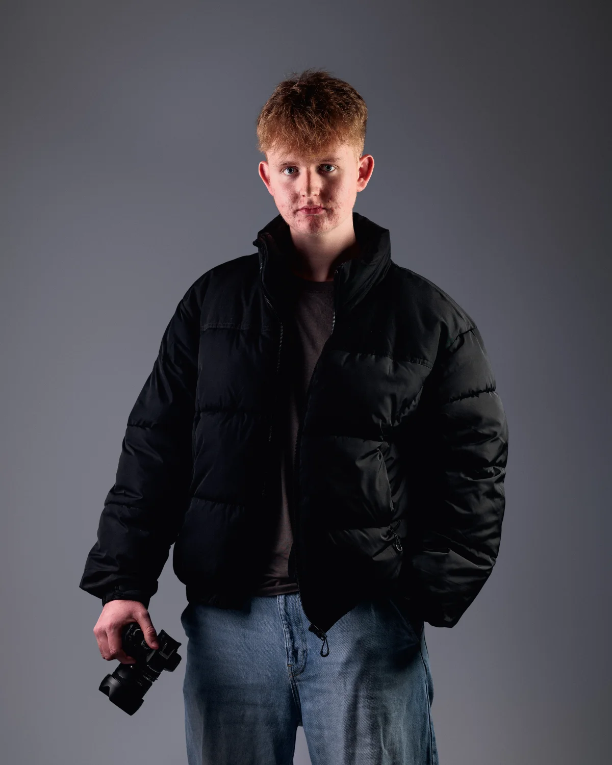 A young man with red hair in a black puffer jacket and blue jeans holding a camera against a grey studio background.