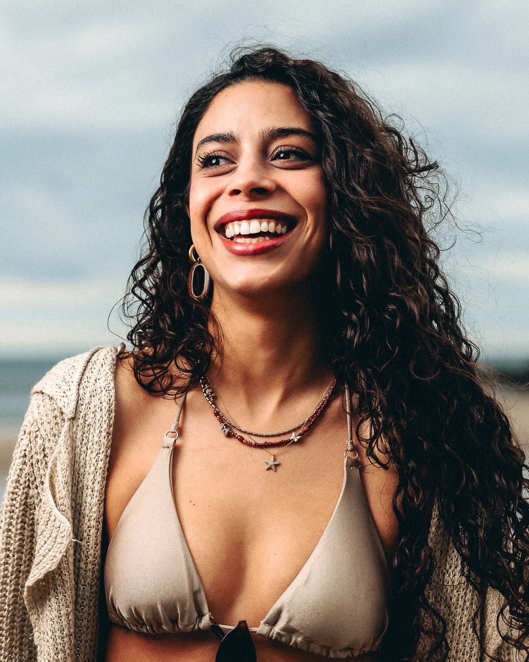 A smiling woman with curly dark hair wearing a beige bikini top and cream cardigan on a beach.