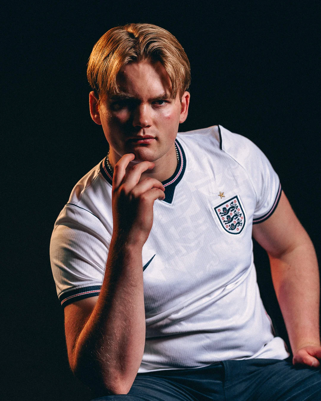 A moody studio portrait of a young man with blond hair wearing an England football jersey, posing thoughtfully.
