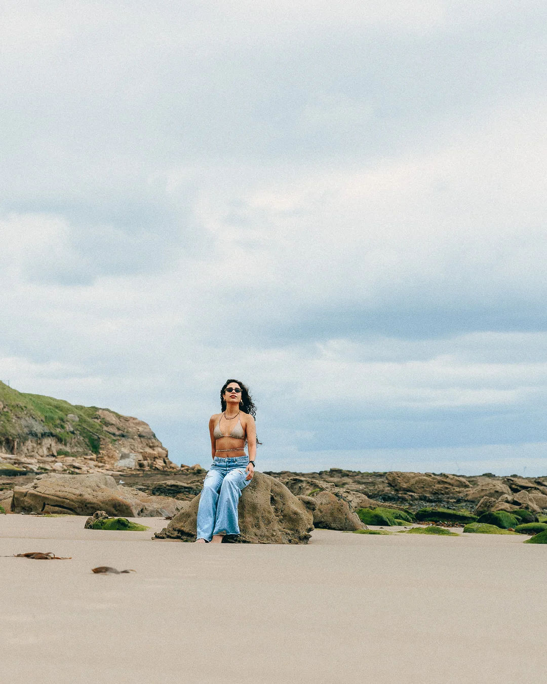 Woman in sunglasses, bikini top and light blue jeans sitting on a rock at a beach under a cloudy sky.