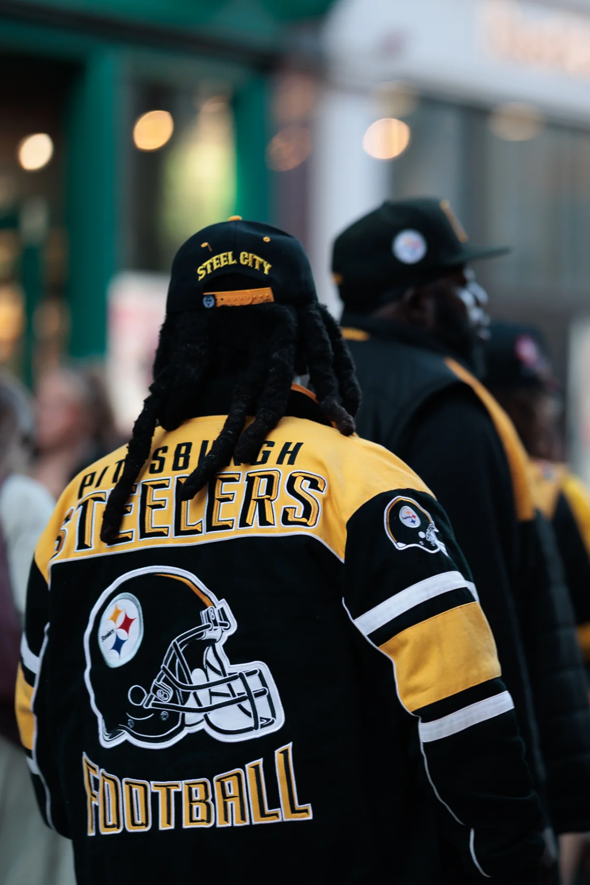 Rear view of a person wearing a Pittsburgh Steelers jacket and 'Steel City' cap in an urban outdoor setting.