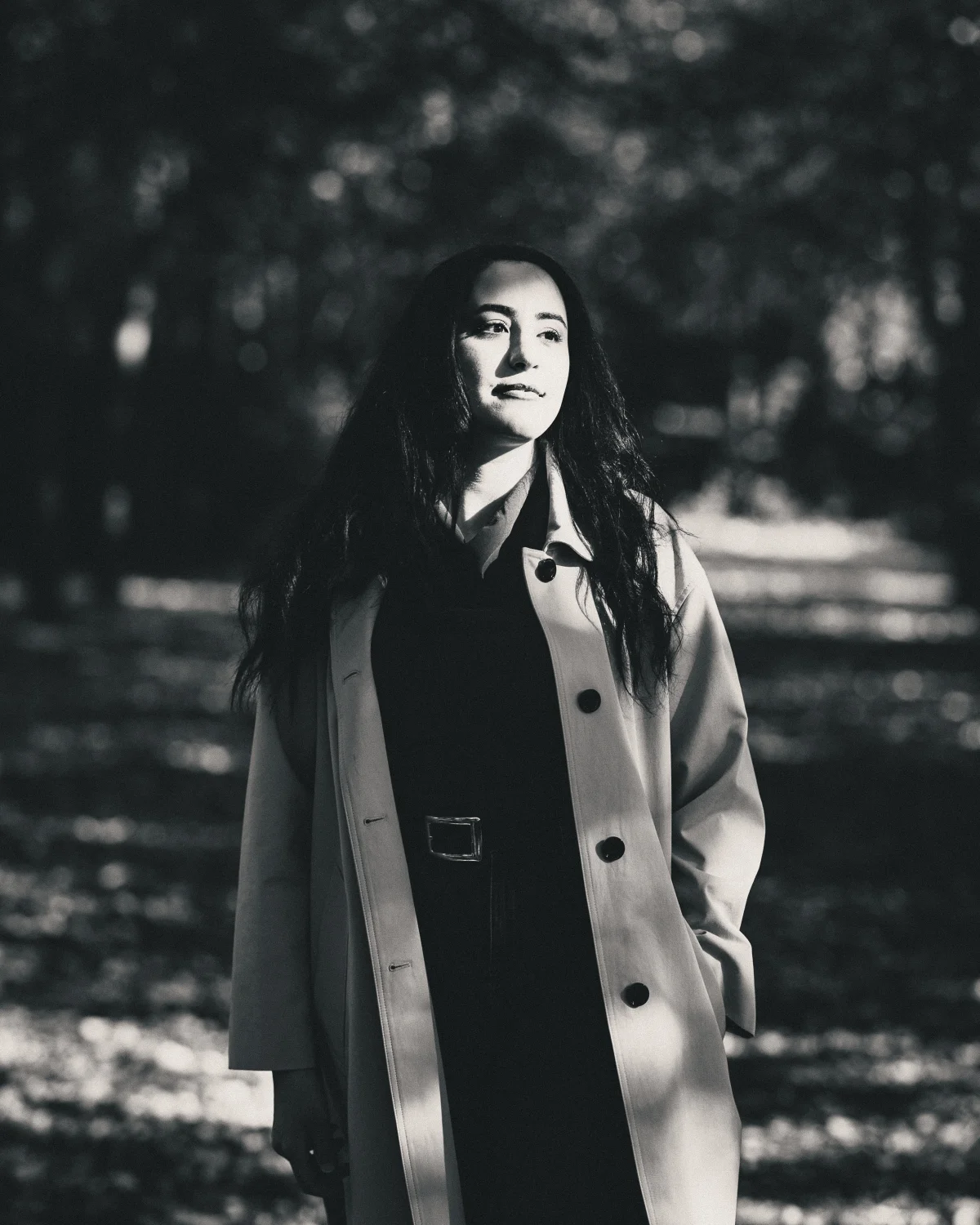 Black and white portrait of a woman in a trench coat posing outdoors in a wooded area.