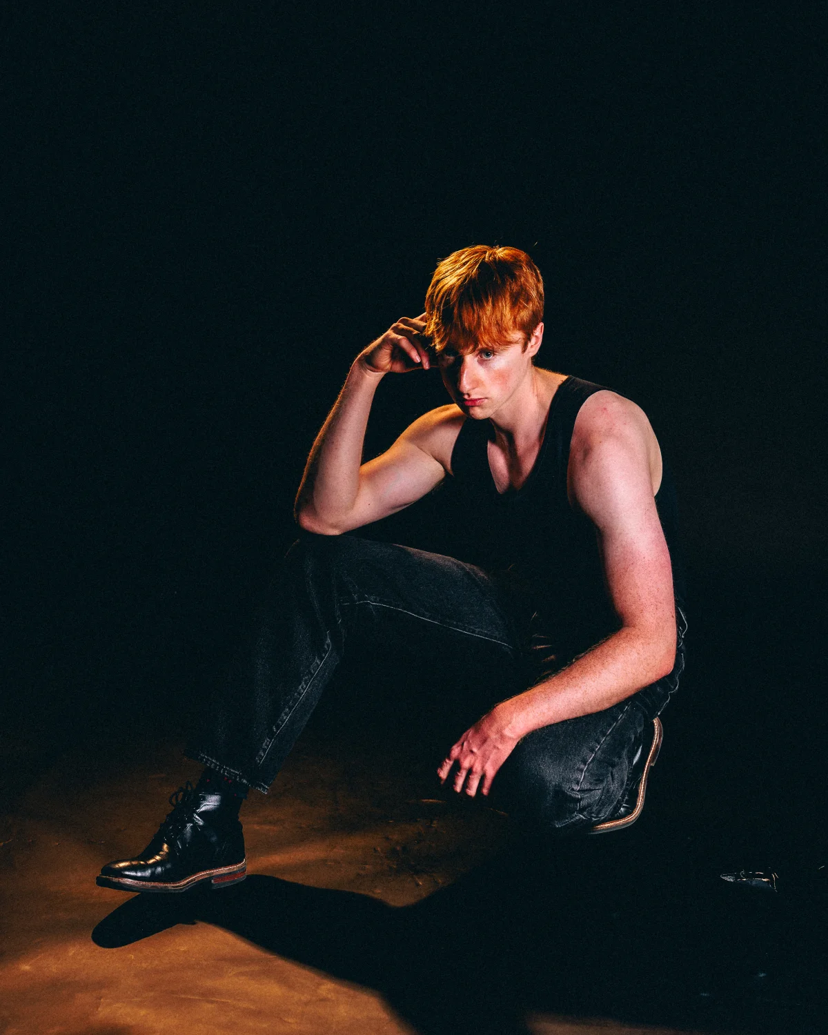 A young man with red hair in a black tank top and jeans posing against a dark background with dramatic lighting.