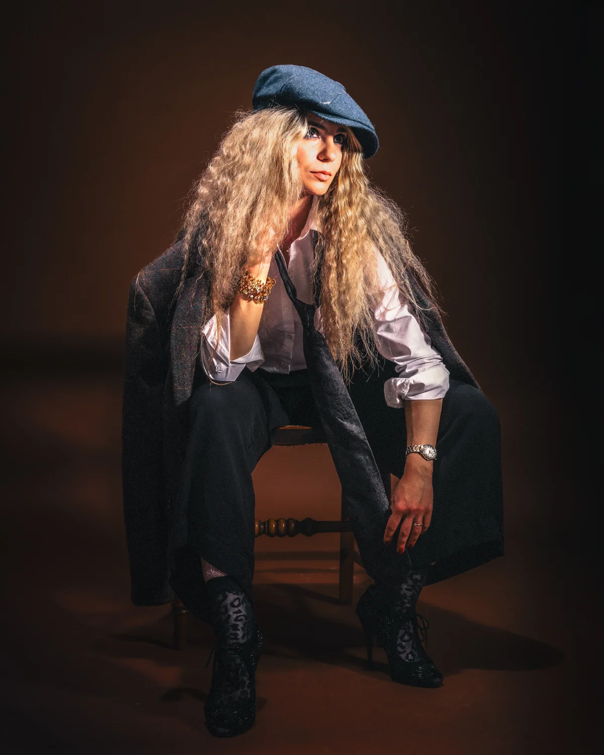 A woman with long curly hair in a blue beret and oversized blazer posing on a chair against a dark background.