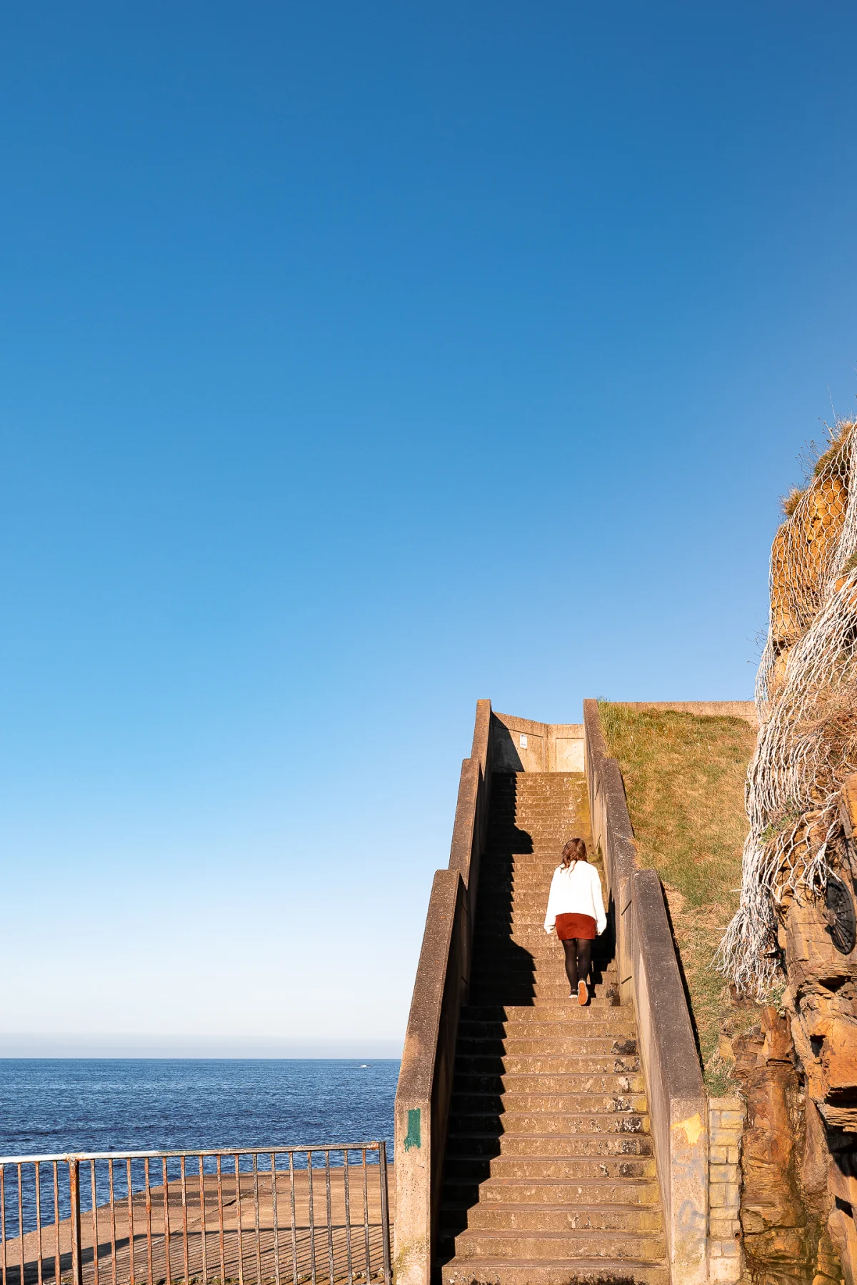 A woman in a white top walking up concrete stairs on a cliff overlooking a blue ocean under a clear sky.