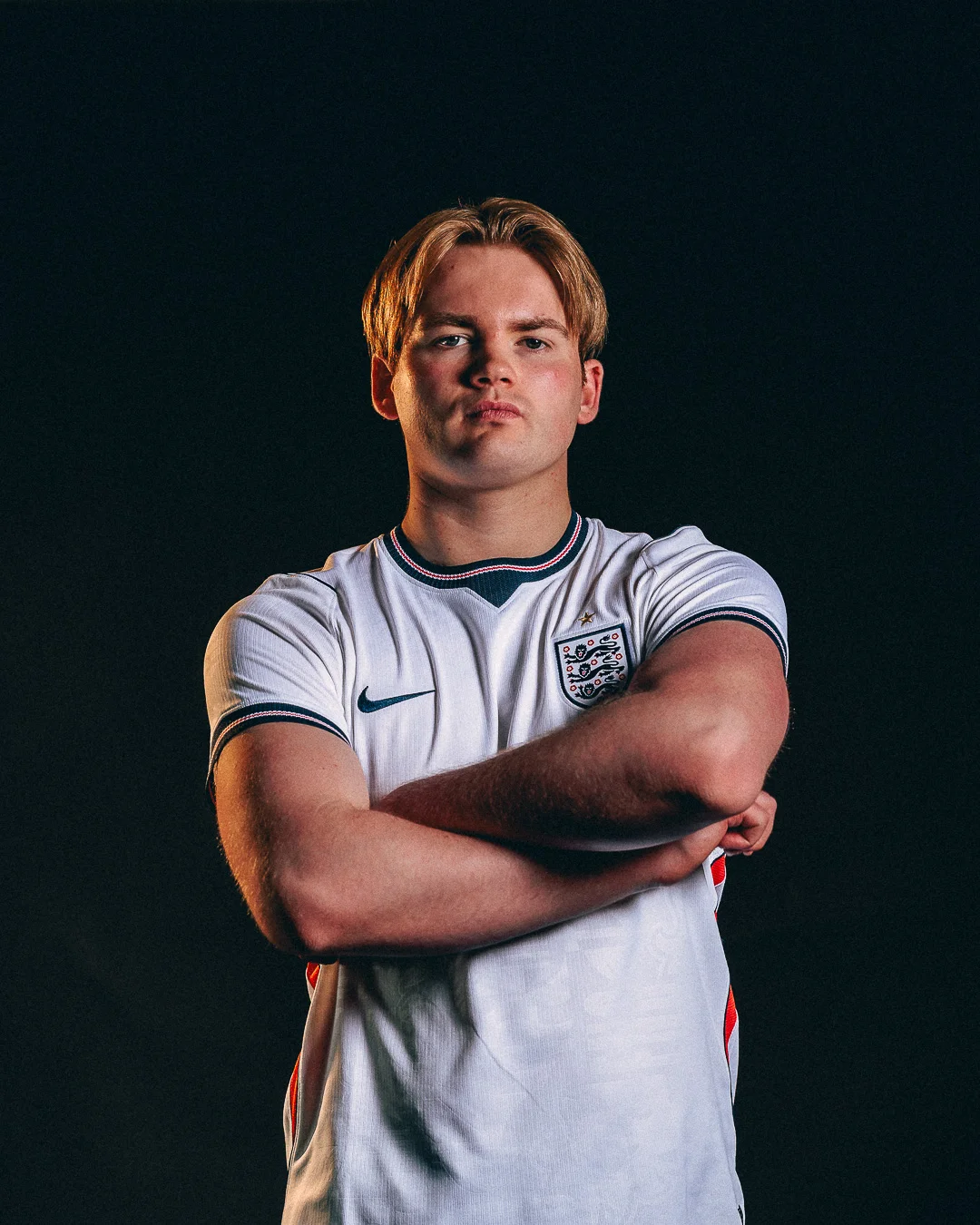 A serious young man with blonde hair wearing an England football jersey, arms crossed against a black background.