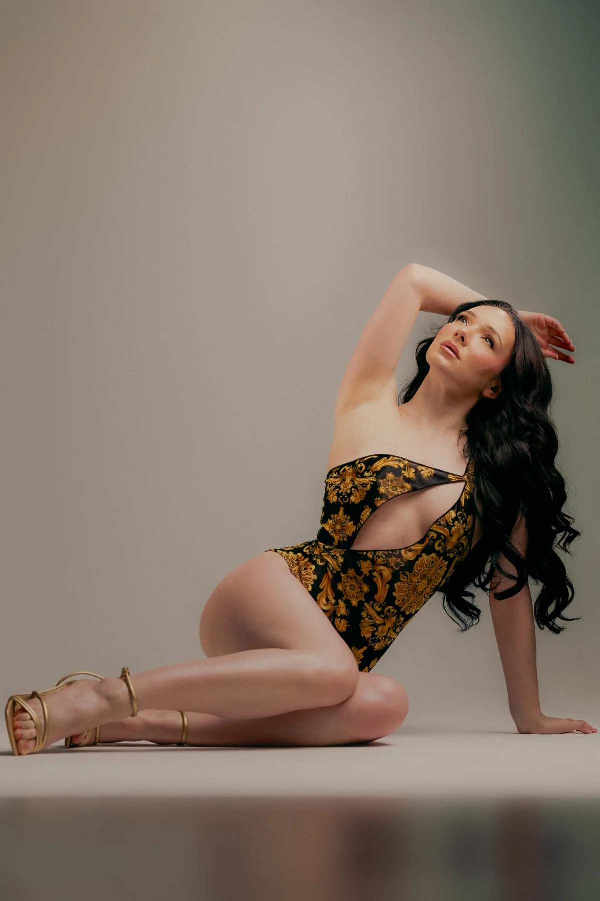 Woman with long dark hair posing in a gold and black patterned cutout bodysuit against a neutral studio background.