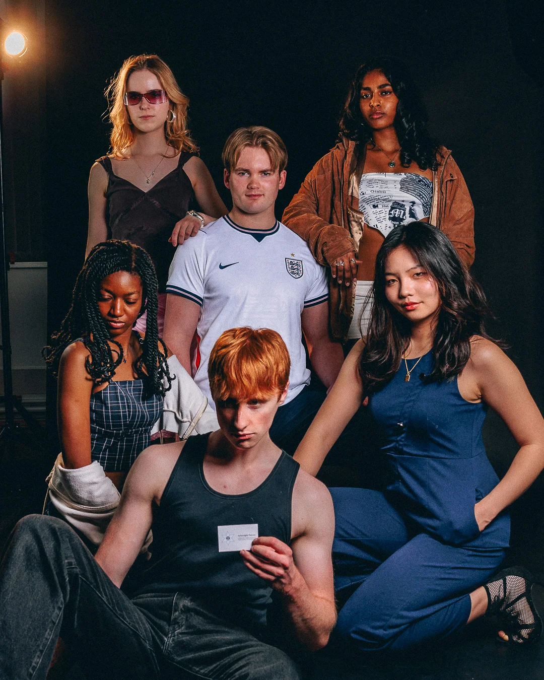 A diverse group of five young adults posing together against a dark background with moody studio lighting.