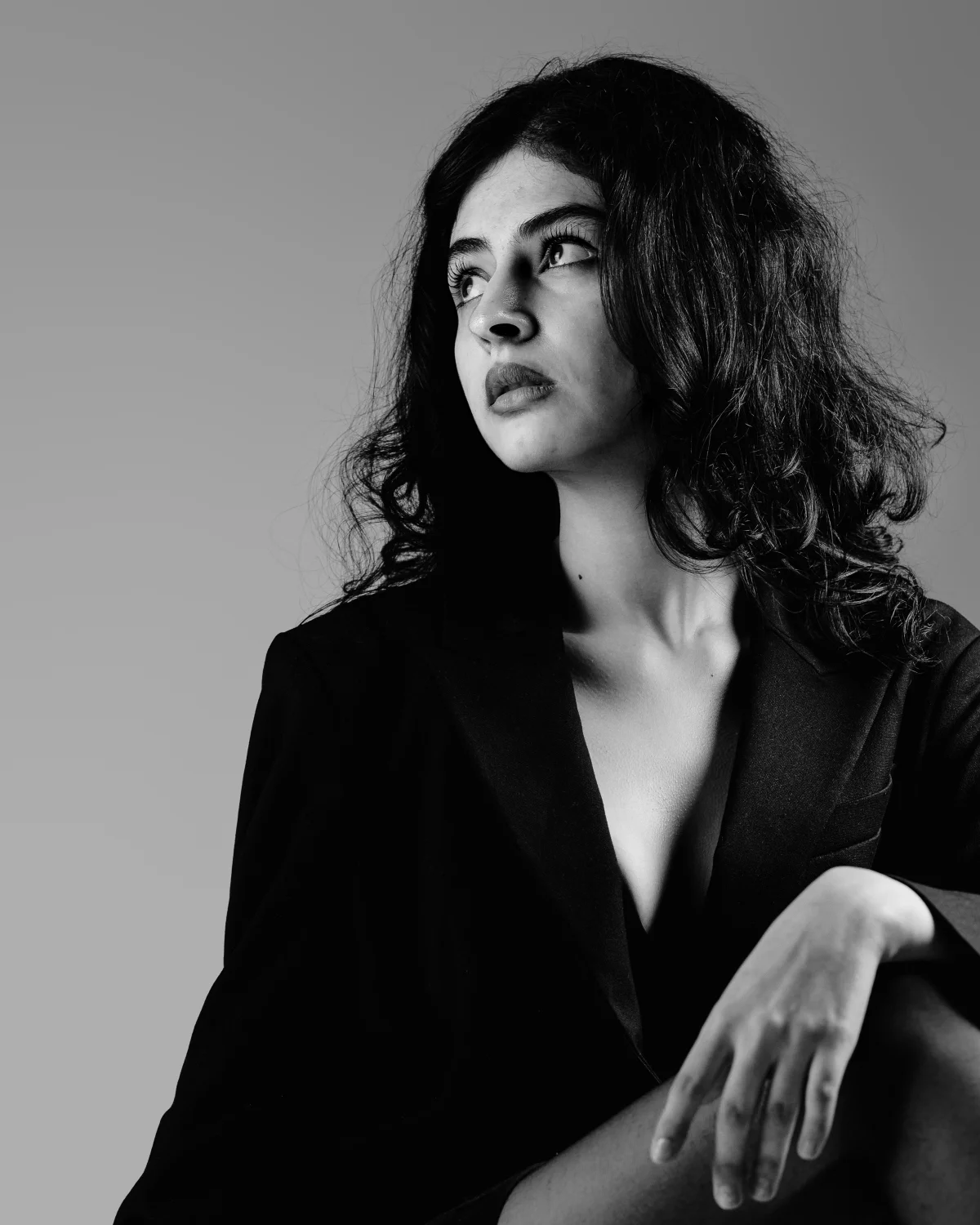 Black and white studio portrait of a woman with curly hair in a dark blazer, looking away thoughtfully.