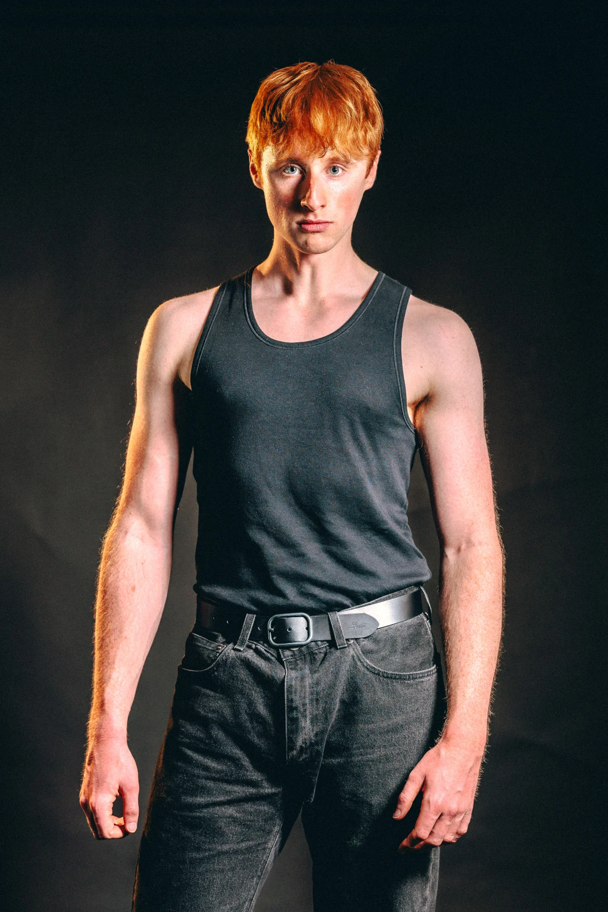 Studio portrait of a red-haired man in a black tank top and dark jeans against a dark background.