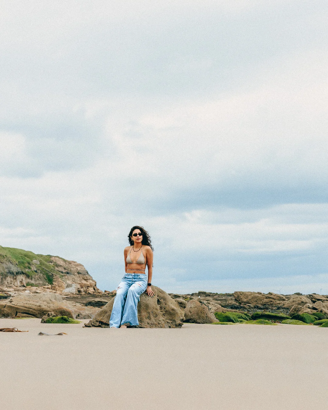 A woman in a bikini top and wide-leg jeans sitting on a rock at a beach under an overcast sky.