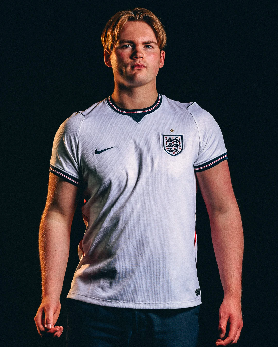 A young man in an England football jersey posing against a black background with dramatic studio lighting.