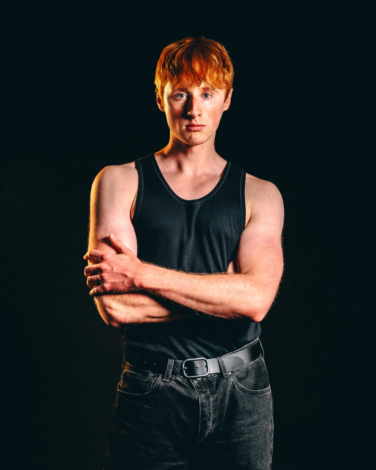 A young man with red hair in a black tank top and jeans posing with crossed arms against a dark background.